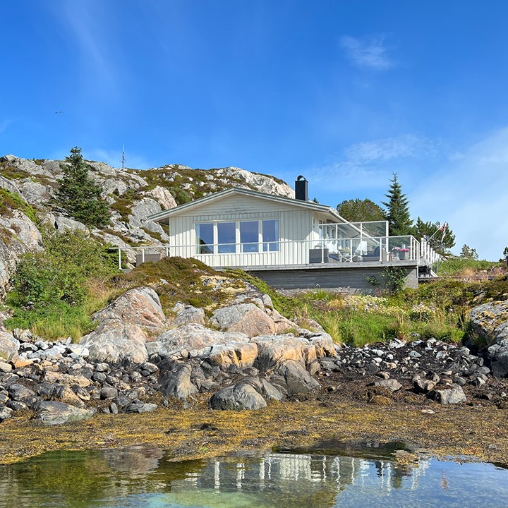 Lysmalt hytte helt nede ved sjøen med svaberg, grønn natur og blå himmel i bakgrunnen. Foto.