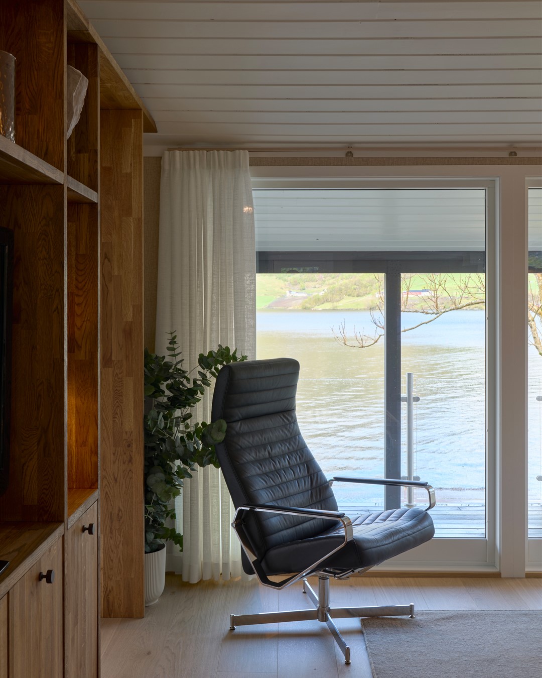 Lys, gulvlang gardin parkert i siden av den store vindusflaten i stuen på Frette-hytta fra Eventyrlig Oppussing. Foran vinduet står en sort lenestol, med bokhylle i tre på siden og utsikt over Stordalsvatnet i bakgrunnen. Foto.