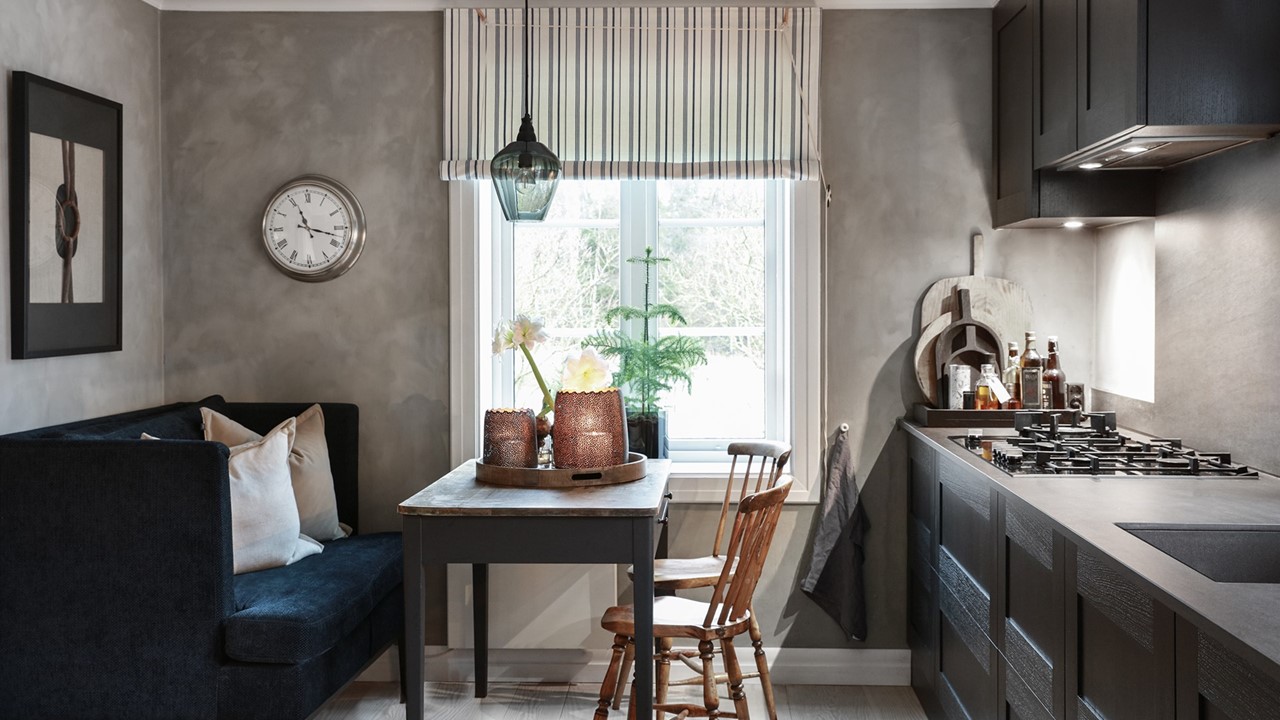 Halvor Bakkes kjøkken med grå vegger, blåstripet 1700-talls roll-up gardin, mørk kjøkkeninnredning, sofa, bord og to stoler. Foto.
