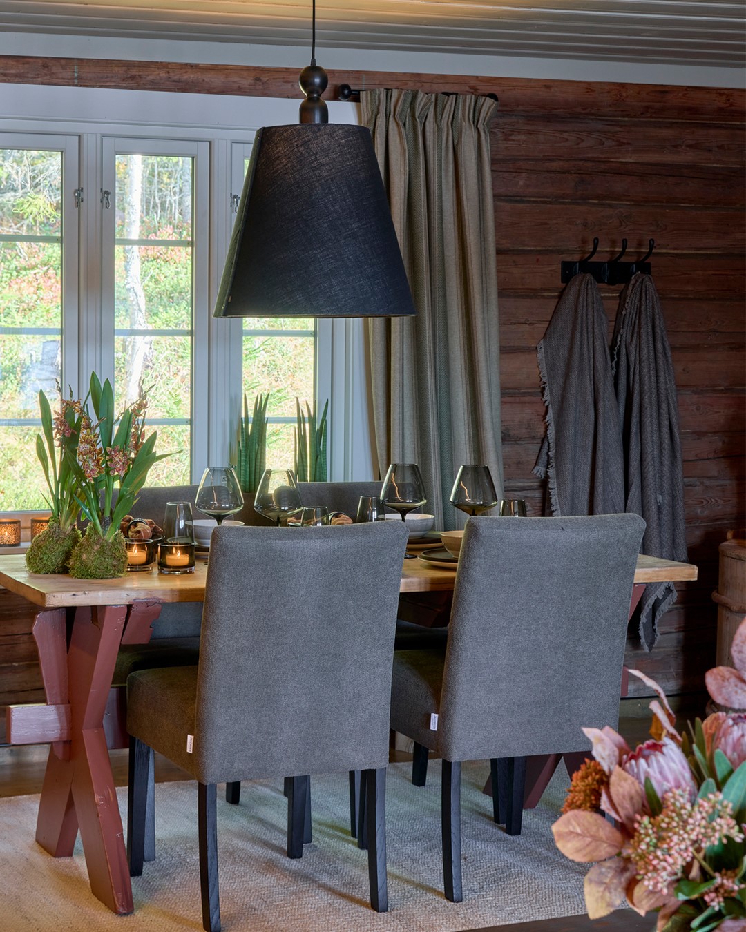 Spiseplass i Mesnali med tømmervegger, dekket bord og lange mønstrede ullgardiner i grønne, beige og burgunder toner, opphengt på svart smijernsstang ved vinduet. Fra Eventyrlig Oppussing Mesnali. Foto.