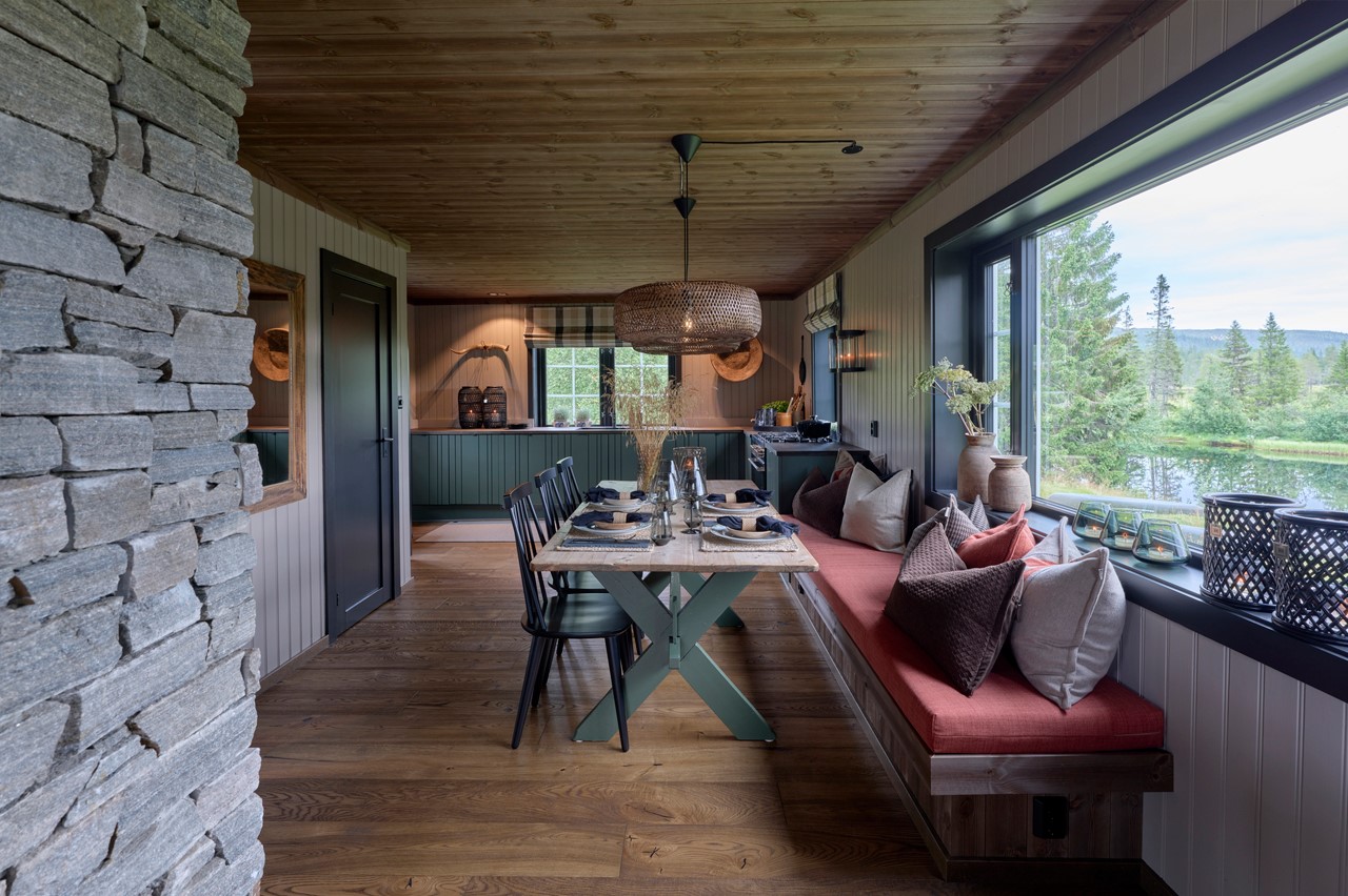 Spisestue og kjøkken fra hytta i Almoseteren. Kjøkkenfronter i grønt. Vindu over kjøkkenbenk med sorte malte lister. Skreddersydd liftgardin i rutete tekstil fra Ian Mankin. Oppdekket spisebord. Sittebenk under vindu med teracotta farget madrass og store puter. Trestoler. Beige vegger og mørkt tregulv og tak. To store flettede taklamper over spisestuen. Fra Eventyrlig Oppussing 2025, Snåsa. Foto. Spisestue og kjøkken fra hytta i Almoseteren. Kjøkkenfronter i grønt. Vindu over kjøkkenbenk med sorte malte lister. Skreddersydd liftgardin i rutete tekstil fra Ian Mankin. Oppdekket spisebord. Sittebenk under vindu med teracotta farget madrass og store puter. Trestoler. Beige vegger og mørkt tregulv og tak. To store flettede taklamper over spisestuen. Fra Eventyrlig Oppussing 2025, Snåsa. Foto.