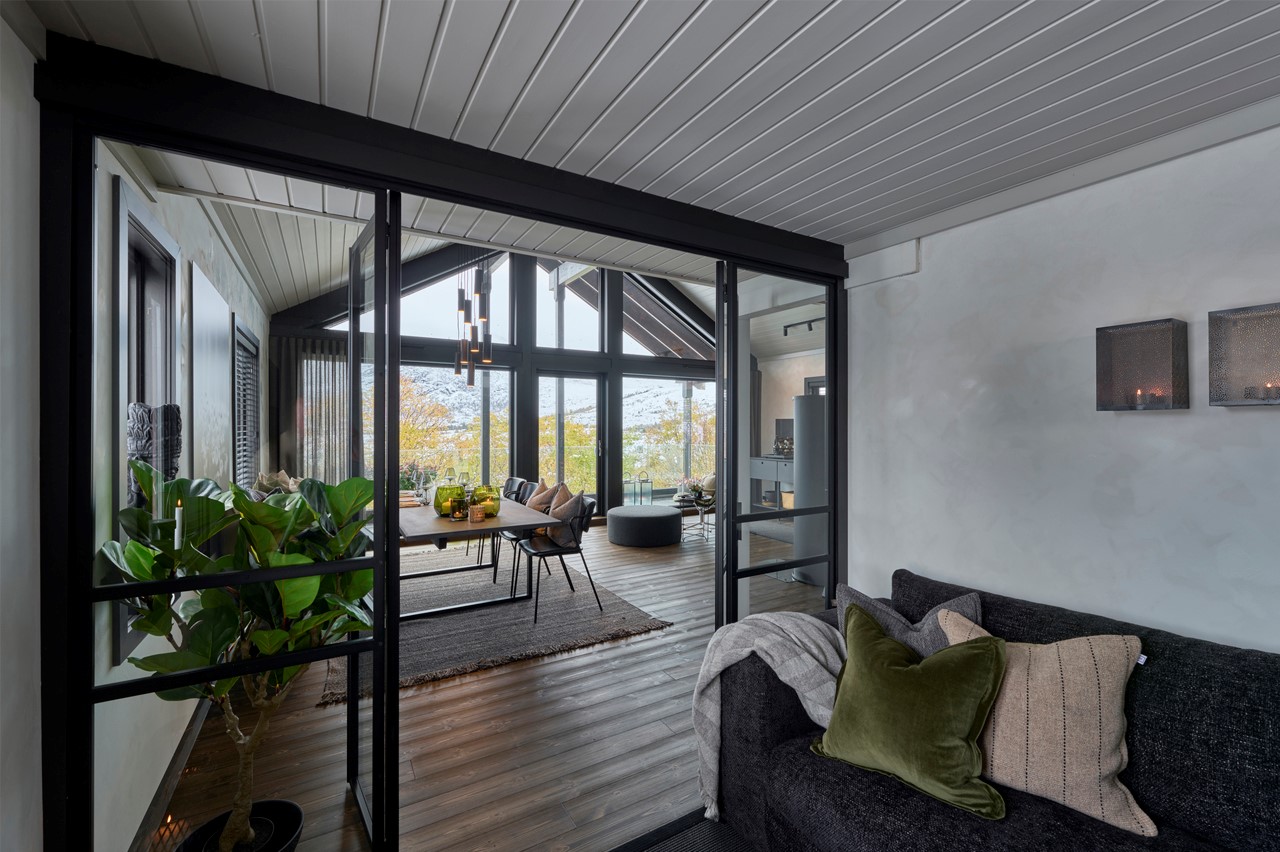 Hovedrommet i hytta. Store vindusflater. Mørkt tregulv. Hvitmalt paneltak. Lys grå kalkmaling på veggene. Vinduer med sorte vinduslister. Spisestue. Store grønne planter. Utsnitt av sofa med store puter i grønt, grå og stripete tekstil. Fra Eventyrlig Oppussing 2025, Sjøholt. Foto. Hovedrommet i hytta. Store vindusflater. Mørkt tregulv. Hvitmalt paneltak. Lys grå kalkmaling på veggene. Vinduer med sorte vinduslister. Spisestue. Store grønne planter. Utsnitt av sofa med store puter i grønt, grå og stripete tekstil. Fra Eventyrlig Oppussing 2025, Sjøholt. Foto.