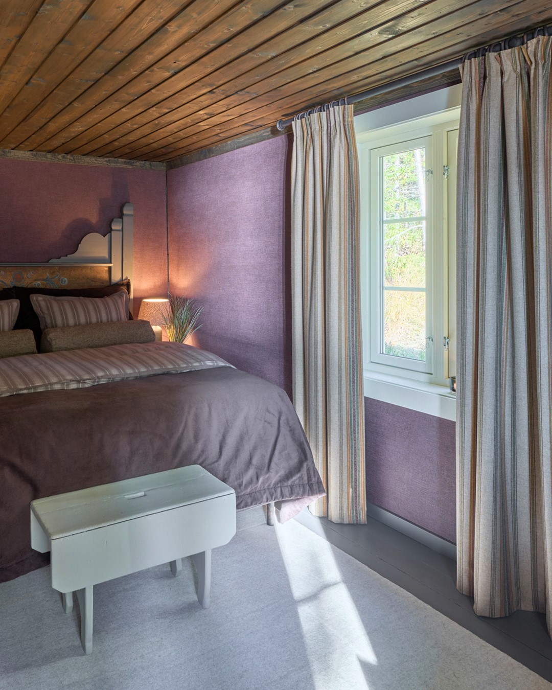 Soverom i Mesnali med lilla vegger, sengetøy i lune toner og lange stripete gardiner som rammer inn vinduet, opphengt på mørk gardinstang. Fra Eventyrlig Oppussing Mesnali. Foto.