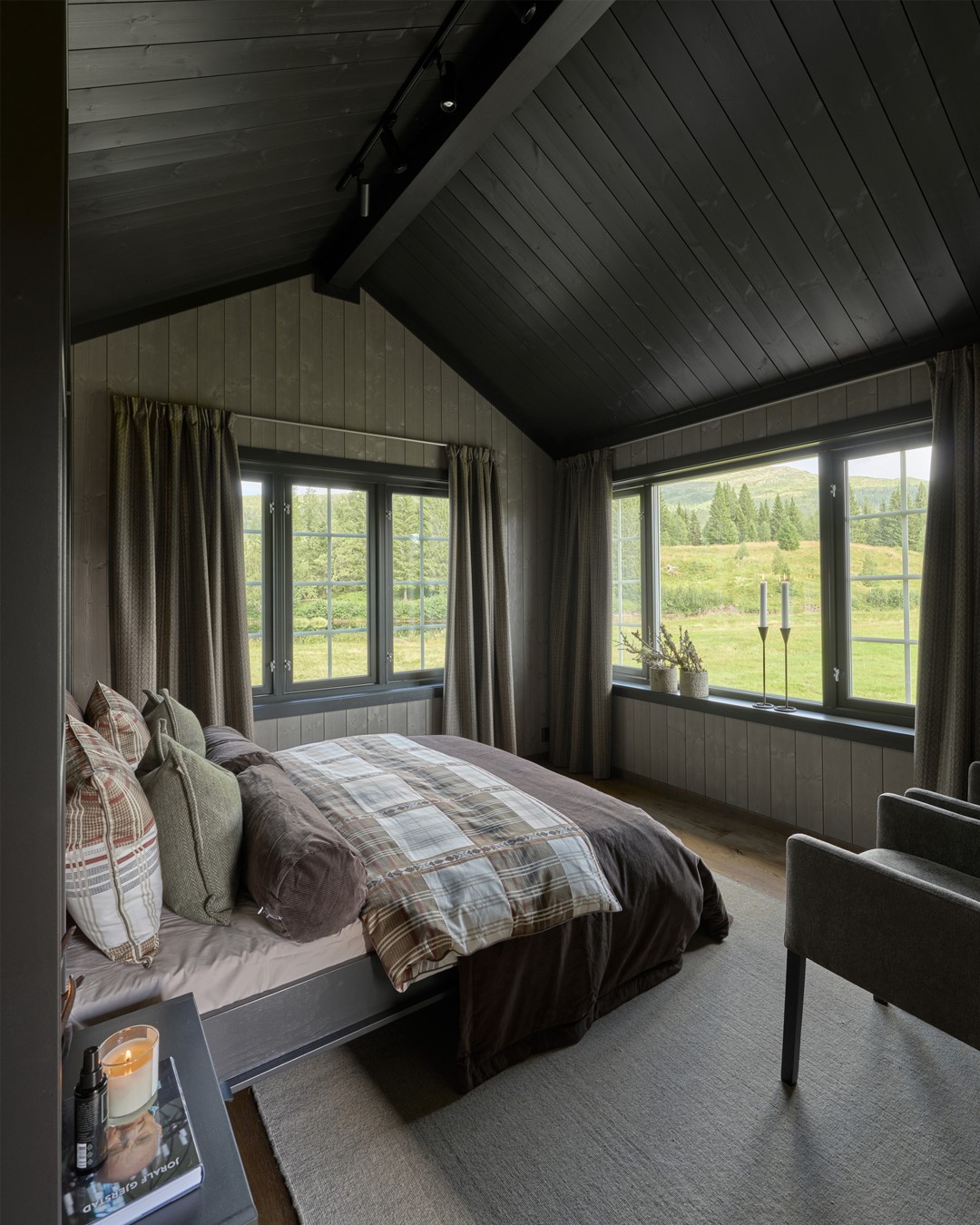 Soverom med oppredd seng, sengetøy og tekstiler i mørk dus lilla, grønne duse farger samt ruter. Lyse panelvegger og sort hylleløsning rundt sengen. Sort tak. Vindu med sortmalte lister. Skreddersydde ullgardiner med lystett fôr sydd i blyantfold. Stort gulvteppe i lys beige. Fra Eventyrlig Oppussing 2025, Almoseteren i Snåsa. Foto. Soverom med oppredd seng, sengetøy og tekstiler i mørk dus lilla, grønne duse farger samt ruter. Lyse panelvegger og sort hylleløsning rundt sengen. Sort tak. Vindu med sortmalte lister. Skreddersydde ullgardiner med lystett fôr sydd i blyantfold. Stort gulvteppe i lys beige. Fra Eventyrlig Oppussing 2025, Almoseteren i Snåsa. Foto.