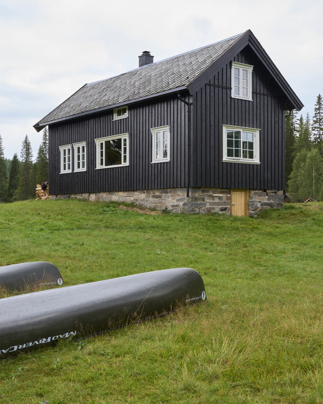 Hytta på Almoseteren. Sortbeiset fasade med hvitmalte vinduskarmer. To kanoer ligger på plenen foran hytta.  Fra Eventyrlig Oppussing 2025, Almoseteren i Snåsa. Foto. Hytta på Almoseteren. Sortbeiset fasade med hvitmalte vinduskarmer. To kanoer ligger på plenen foran hytta.  Fra Eventyrlig Oppussing 2025, Almoseteren i Snåsa. Foto.