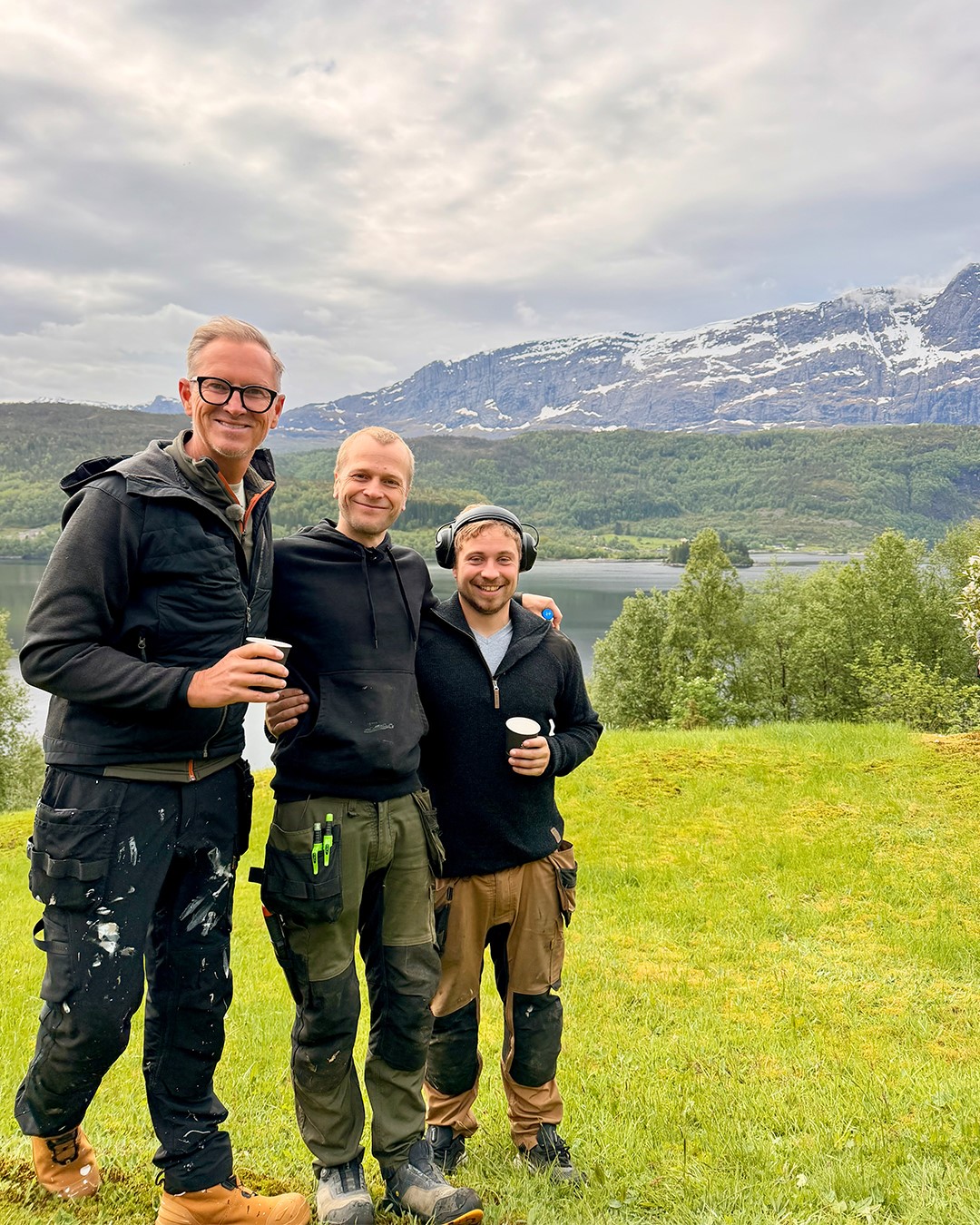 Halvor Bakke & Co står samlet på en grønn gresslette med fjord, skogkledde åser og høye fjell med snøflekker i bakgrunnen. Det vestlandske landskapet rundt Naustdal danner en dramatisk ramme rundt bildet. Fra Eventyrlig Oppussing Naustdal. Foto.