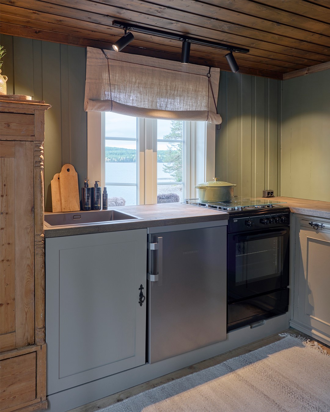 Kjøkken i Mesnali med grønne panelvegger, trebenkeplate og utsikt mot vannet, innrammet av en skreddersydd, naturfarget 1700-talls liftgardin med synlig snorsystem. Fra Eventyrlig Oppussing Mesnali. Foto.