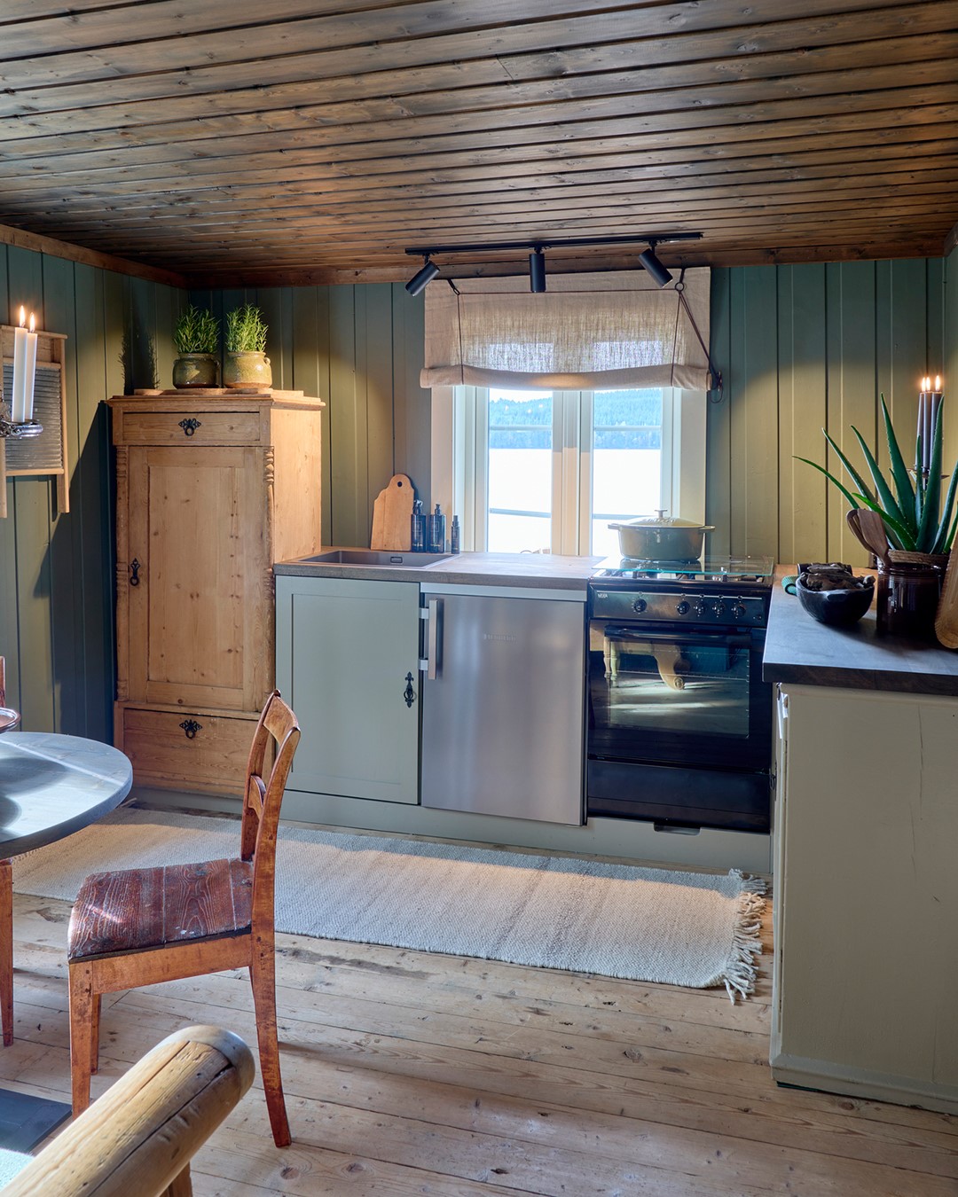Rustikt kjøkken i Mesnali med grønne panelvegger, tregulv og lys liftgardin i naturfarget tekstil over kjøkkenvinduet. Fra Eventyrlig Oppussing Mesnali. Foto.