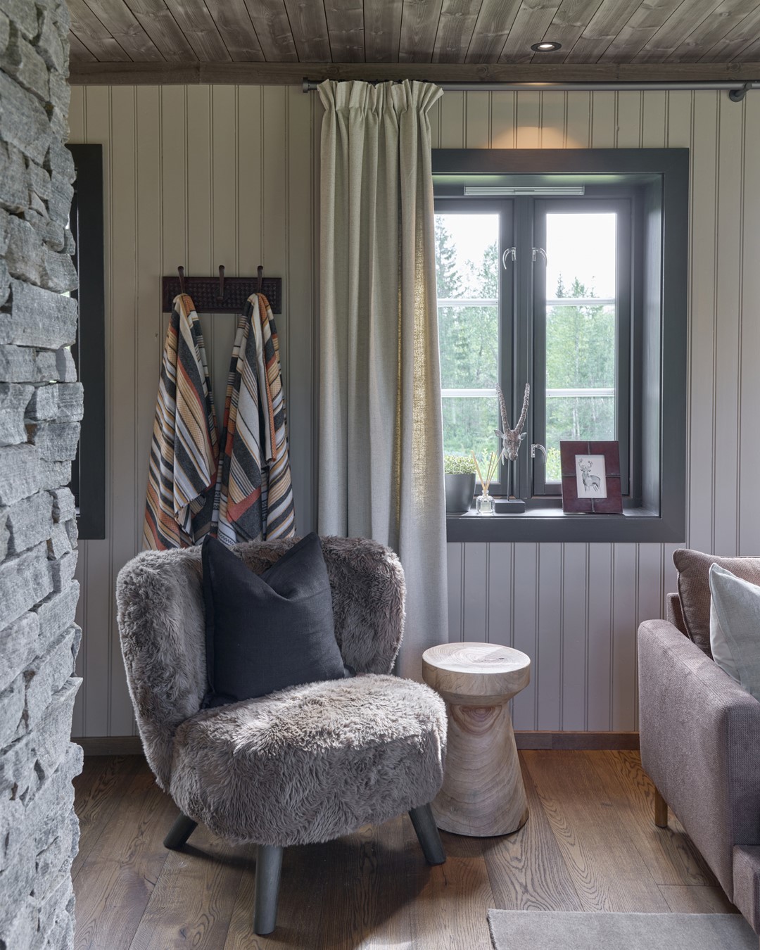 Lesekrok ved vinduet i stua. Stor myk lesestol i teddytekstil. Varmt tregulv og tak. Lyse vegger. Vindu med sortemalte lister. Skreddersydde gardiner i blyantfold i lys beige tekstil fra Roma. Store gode pledd i stripete tekstil henger på kroker bak stolen. Fra Eventyrlig Oppussing 2025, Almoseteren i Snåsa. Foto. Lesekrok ved vinduet i stua. Stor myk lesestol i teddytekstil. Varmt tregulv og tak. Lyse vegger. Vindu med sortemalte lister. Skreddersydde gardiner i blyantfold i lys beige tekstil fra Roma. Store gode pledd i stripete tekstil henger på kroker bak stolen. Fra Eventyrlig Oppussing 2025, Almoseteren i Snåsa. Foto.