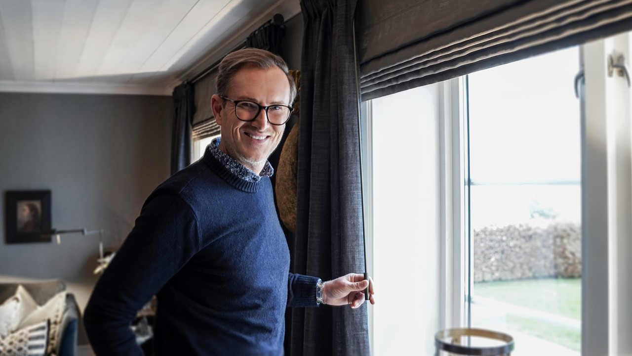 Halvor Bakke står ved vinduet i stuen sin med mørke gardiner og liftgardiner. Foto.