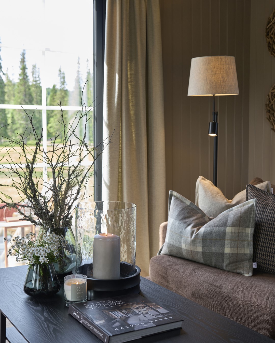 Utsnitt av stue og sofakrok med skreddersydd pute i rutete ull-tekstil. Skreddersydde gardiner i blyantfold i lys beige, tekstilen Kyri fra Roma, i fargen Taupe. Sort sofabord med blomster, kvister og lyslykter. Fra Eventyrlig Oppussing 2025, Almoseteren i Snåsa. Foto. Utsnitt av stue og sofakrok med skreddersydd pute i rutete ull-tekstil. Skreddersydde gardiner i blyantfold i lys beige, tekstilen Kyri fra Roma, i fargen Taupe. Sort sofabord med blomster, kvister og lyslykter. Fra Eventyrlig Oppussing 2025, Almoseteren i Snåsa. Foto.