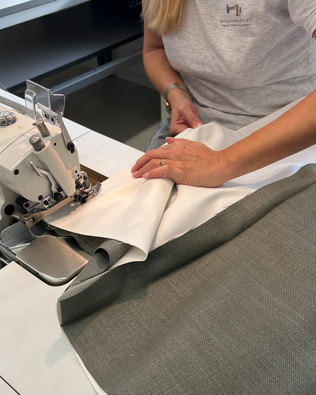 Fra sømstudio ser vi en symaskin, tekstil med for og hendene til sømmerske som syr liftgardin. Foto.