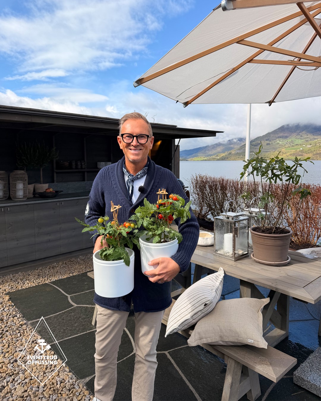 Halvor Bakke på uteplassen ved Frette-hytta i Eventyrlig Oppussing, med tomatplanter i hendene, utekjøkken og utsikt mot vann og fjell. Foto.