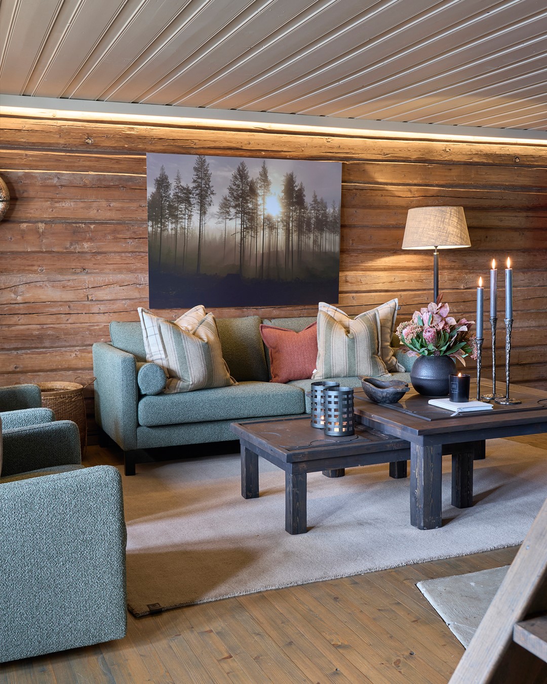 Stue i Mesnali med tømmervegger, grønn sofa og sofabord, der putene i mønstret ulltekstil tar opp igjen stoff og farger fra gardinløsningen i rommet. Fra Eventyrlig Oppussing Mesnali. Foto.