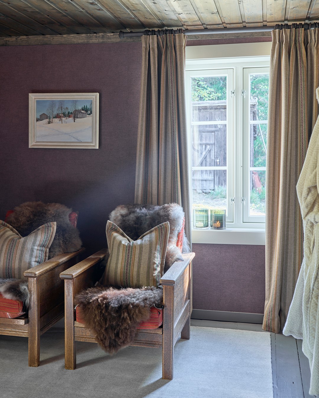 Sitteplass på soverommet i Mesnali med trestoler, saueskinn og puter, mens lange stripete ullgardiner i beige, grønt og burgunder rammer inn vinduet på mørk gardinstang. Fra Eventyrlig Oppussing Mesnali. Foto.