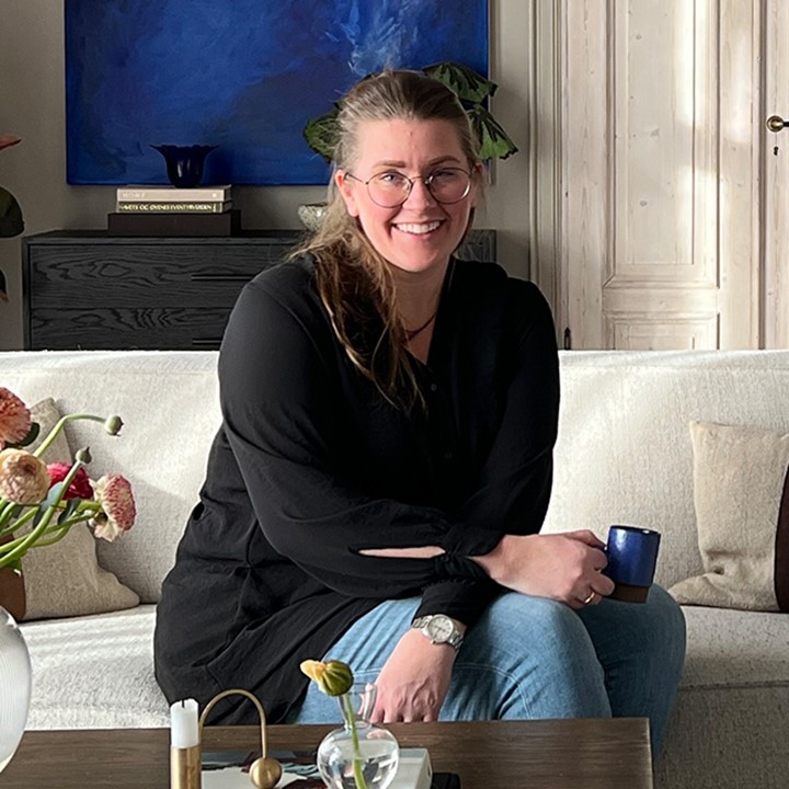 Interiørdesigner og trendforsker Kristin Lindhjem sitter i lys sofa i stue med lyse vegger og lingardiner fra tak til gulv. Gamle doble dører og blått kunstverk på veggen. Foto.