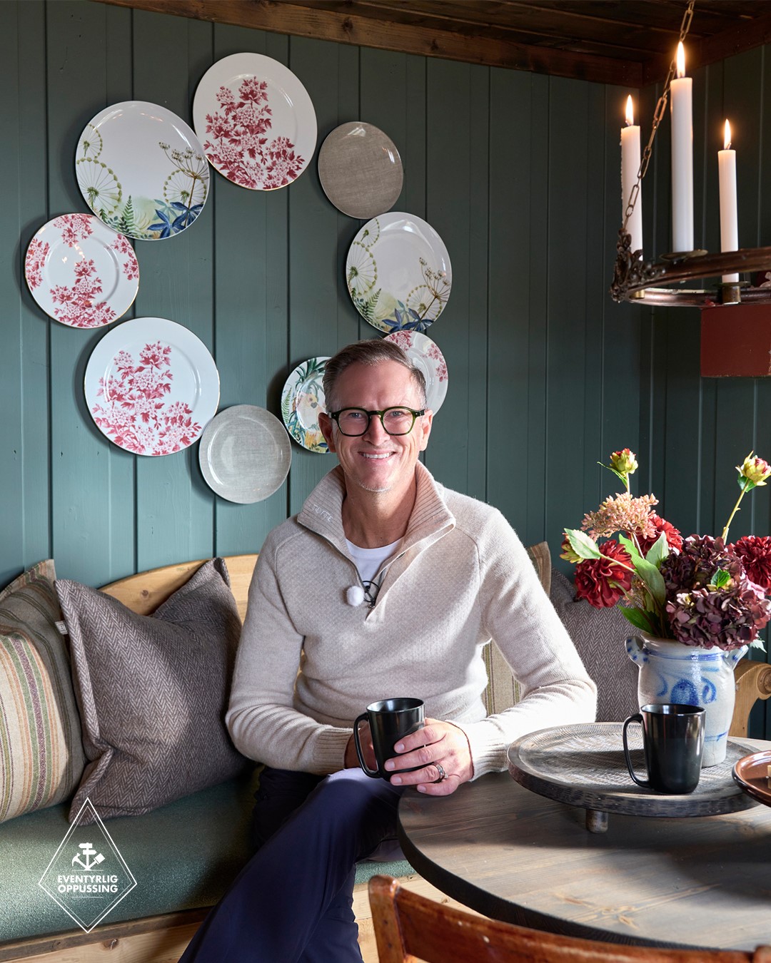 Halvor Bakke sitter ved spiseplassen i hytta i Mesnali, omgitt av mørk grønn panelvegg, trebenk med puter, blomster og rustikke interiørdetaljer. Fra Eventyrlig Oppussing Mesnali. Foto.